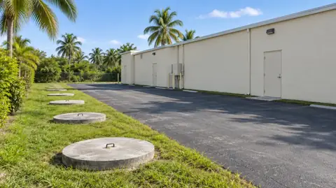 A row of large round concrete septic lids in a grass strip beside the asphalt parking area of a cream commercial building