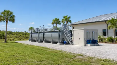 A long grey packaged wastewater treatment plant with walkways and blowers on a gravel pad beside a single-story building