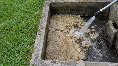A jet of water from a hose nozzle breaking up a thick tan scum mat inside an open concrete septic tank